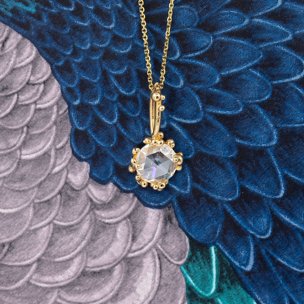 Close-up of a yellow gold pendant necklace featuring a rose-cut moissanite framed by organic gold bead details, displayed against a blue and grey feather-patterned fabric background.