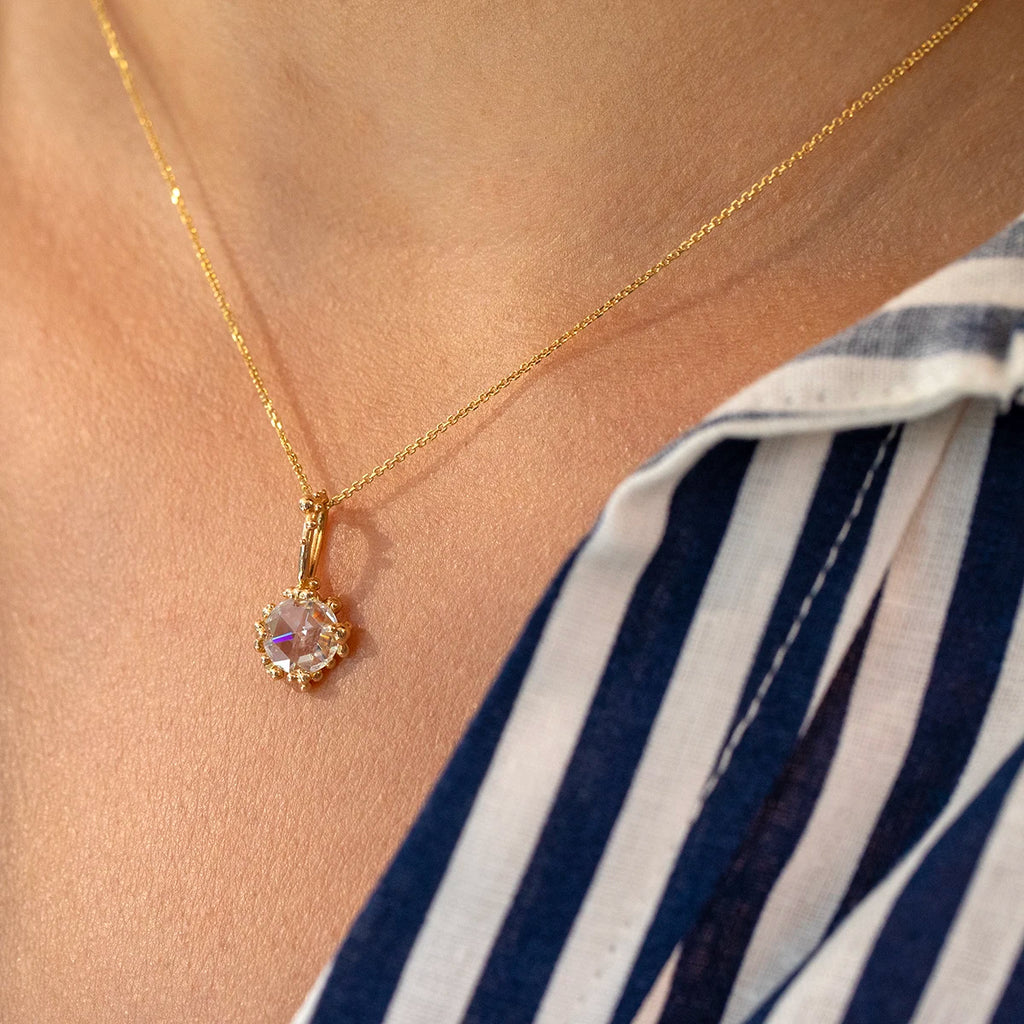 Close-up of a woman wearing a yellow gold necklace with a rose-cut moissanite pendant framed by small gold bead details, styled with a blue-and-white striped shirt.