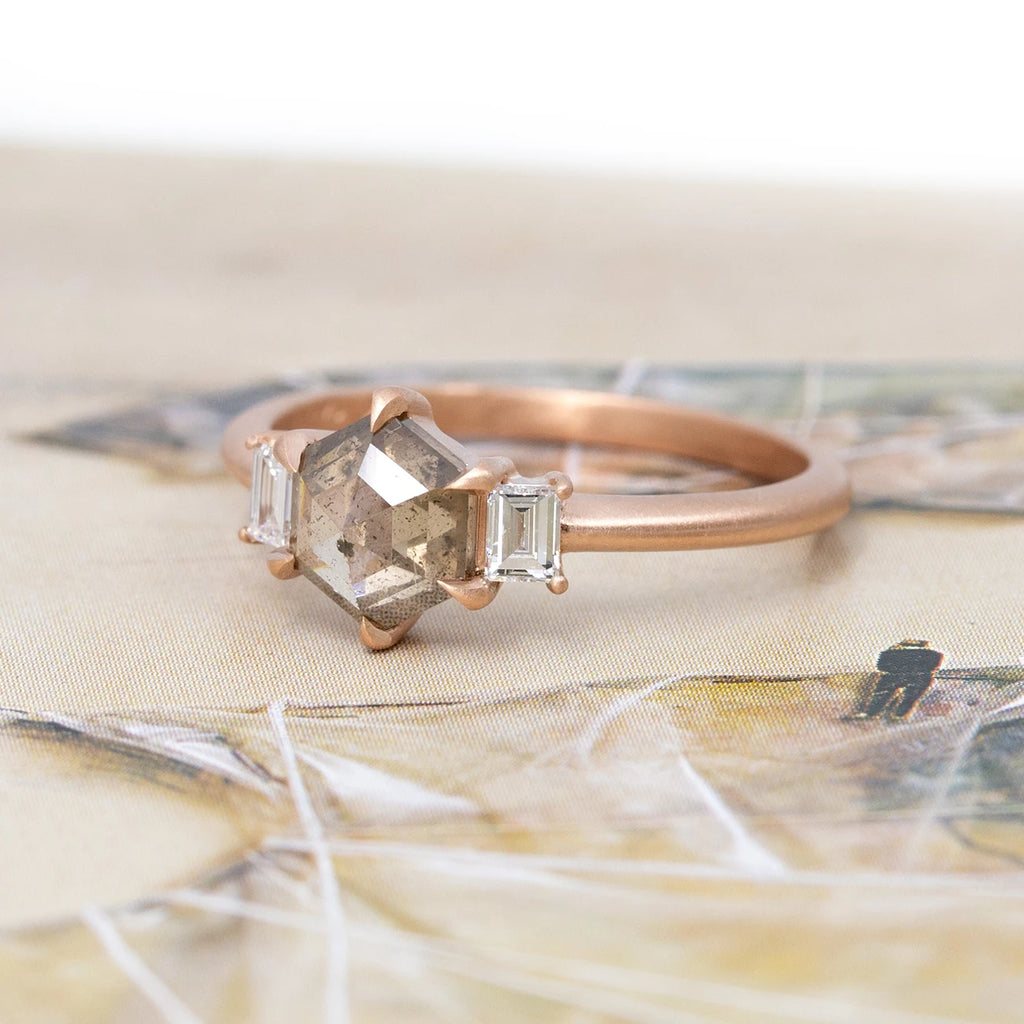 Side view of a classic rose gold engagement ring with a hexagonal champagne diamond and two baguette-cut white diamonds, set with eagle claws, photographed against an abstract artistic background. Made with Fairmined-certified gold by Ottawa-based independent brand Fluid Jewellery. A fine jewelry piece available at Ruby Mardi, a jewelry store specializing in unique designer jewelry, as Gem Breakfast does.