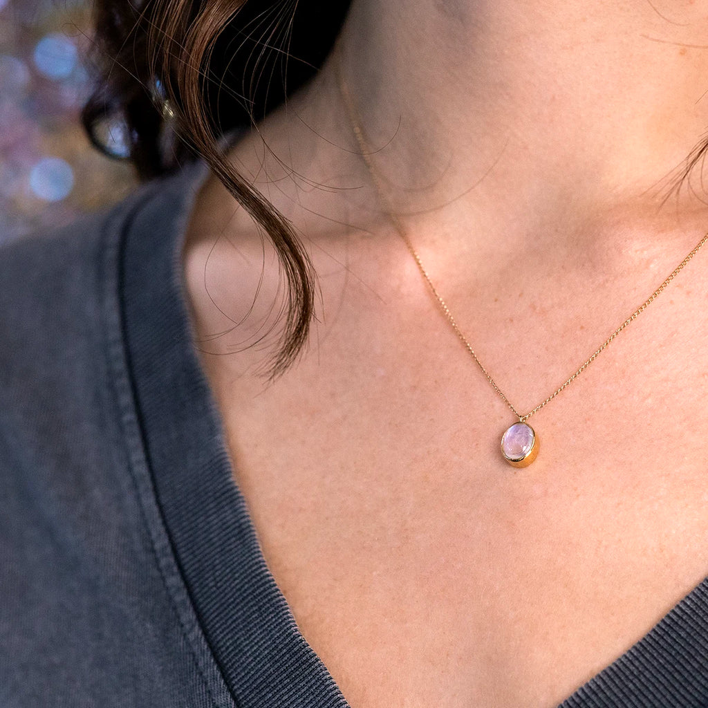 Rose quartz pendant in yellow gold worn by a woman with a grey V-neck t-shirt, set against a vibrant multi-colored sparkle background. A delicate and elegant accessory available at Ruby Mardi.