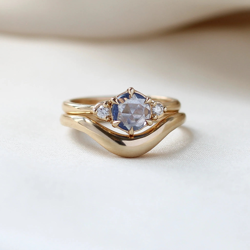 Rose-cut round blue sapphire engagement ring framed by brilliant diamonds, handcrafted in 14k yellow gold by Yuliya Chorna, shown at Ruby Mardi Montreal. Seen stacked with a large curved gold band.
