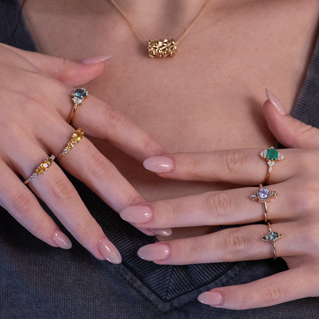 A beautiful portrait of a woman with her hands gently resting on her heart, showcasing exquisite designer jewelry. She wears a gray t-shirt that highlights a textured gold tube pendant, complemented by six unique rings, each telling its own story. These exceptional pieces, featuring natural sapphires, emeralds, and both black and white diamonds, celebrate the talent of Canadian designers and add a touch of elegance to her style. A true statement of passion for fine craftsmanship.
