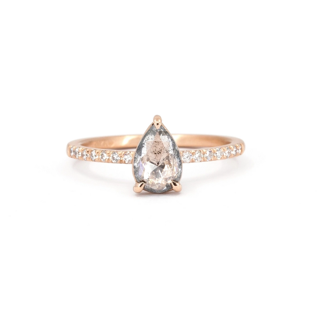 Classic rose gold solitaire engagement ring with a central pear salt and pepper diamond set with eagle claws, photographed on a white background. The band of this bridal ring is set with small round brilliant diamonds in half-eternity. Made with Fairmined-certified gold by Ottawa-based independent brand Fluid Jewellery. A fine jewelry piece available at Ruby Mardi, a jewelry store specializing in unique designer jewelry, as Gem Breakfast does.