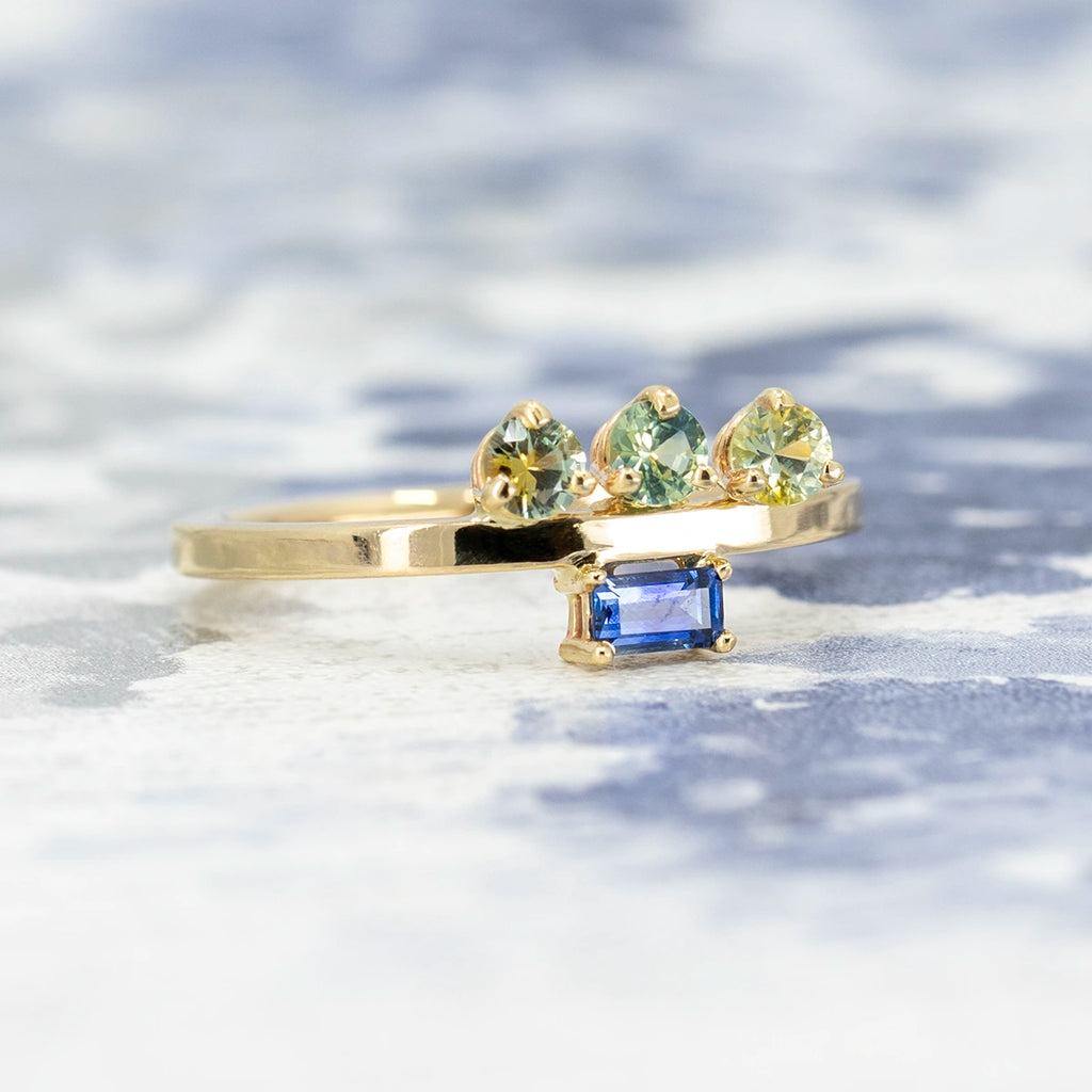 A close up of an original fine jewelry piece photographed on an abstract blue background. The rings features a flat 14k yellow gold recycled band. One one side of it we can see 3 bicolor sapphires of a round shape (blue and yellow), and on the other side a royal blue baguette sapphire. This unique engagement ring can be worn either sides. It's a design from In the Light of Day Jewelry, and it is available in Montreal, Canada, at indie jewelry store Ruby Mardi.
