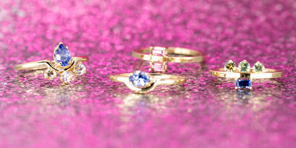 Banner photo presenting a collection of 4 gold rings from an independent jeweller. All the rings are one-of-a-kind and features sapphires. The jewels are photographed on a pink sequin background.