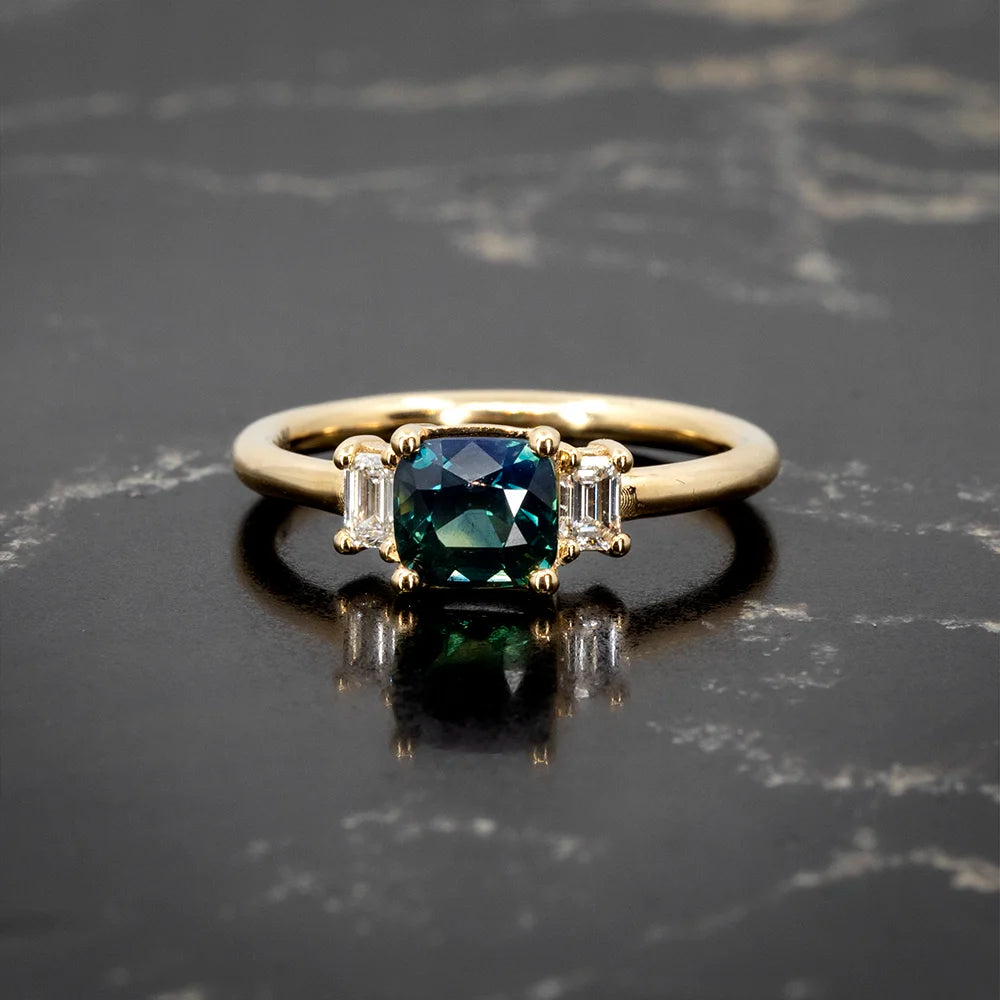 Teal sapphire engagement ring by Sheena Purcell, featuring a cushion-cut center stone flanked by baguette diamonds, set in yellow gold. Available at Ruby Mardi, Montreal.