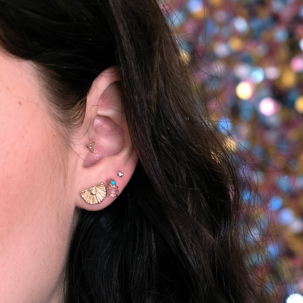 Close-up of an ear styled with a unique mix of earrings: a gold fan-shaped stud with a diamond, a delicate pink tourmaline and turquoise accent earring, and minimalist gold and diamond studs. Set against dark hair and a colorful, sparkling backdrop.