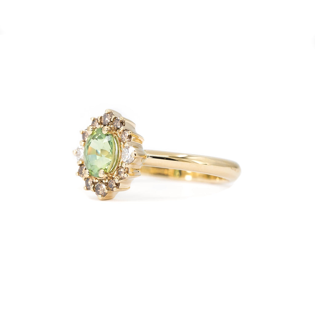 ide view of a handcrafted yellow gold engagement ring featuring a vivid round green sapphire at the center, surrounded by a halo of champagne diamonds with two white diamond accents, designed by Ruby Mardi Jewelry in Montreal.