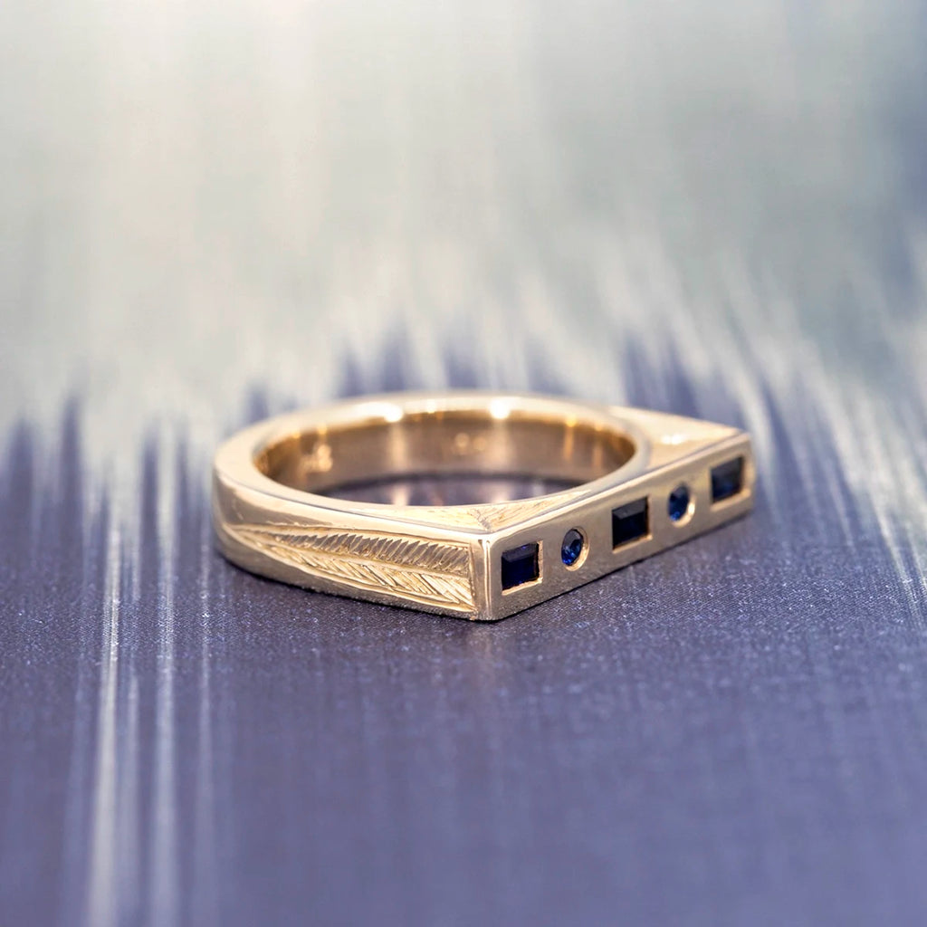 Side view of a hand-engraved 14k yellow gold geometric ring set with dark blue baguette and round sapphires, Rebel & Rogue Jewelry, photographed on a textured blue background for Ruby Mardi Montreal.