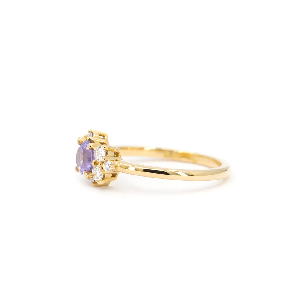 Side view of a gold ring handcrafted by Justine Quintal, featuring a round purple sapphire center stone flanked by asymmetrically arranged small round white diamonds, displayed on a white background.