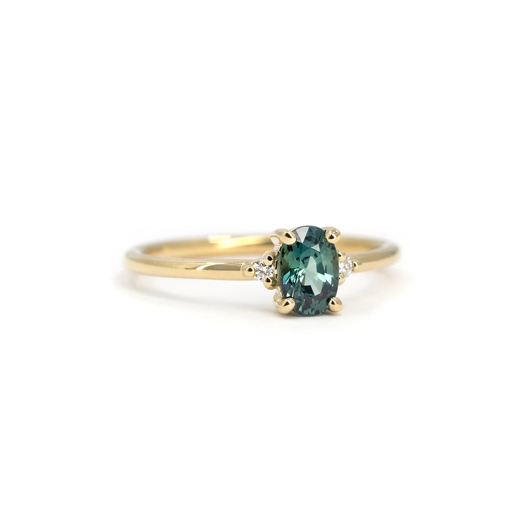 Teal oval sapphire engagement ring with diamond side stones, set in yellow gold. Handmade by Maksym Jewelry in Montreal. Available at Ruby Mardi.