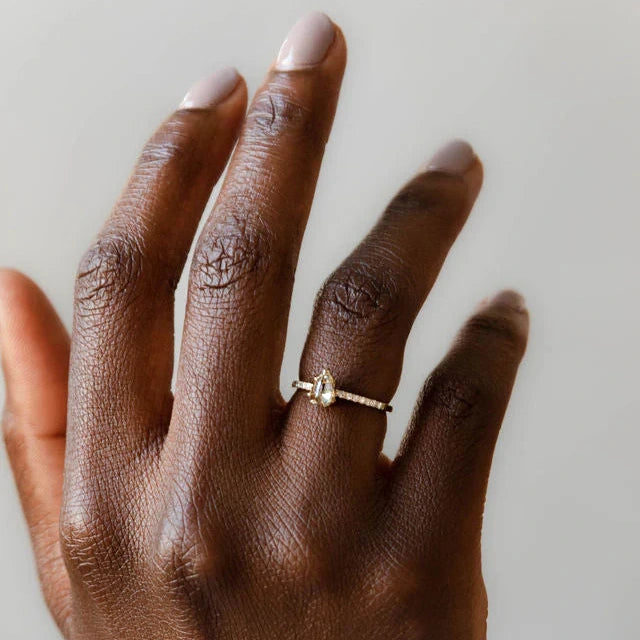 A solitaire engagement ring is seen in a close-up on a black woman's hand. The ring is set in a half eternity, and the central diamond is shield-shaped. This jewel was handcrafted in Canada from Fairmined-certified ethical gold. It is available at independent jewelry store Ruby Mardi, located in Montreal's Little Italy. 