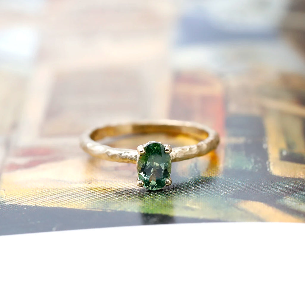 Rough gold band solitaire engagement ring showing an oval green sapphire showing hues of yellow. This one-of-a-kind was handmade in Toronto by Anouk Jewelry and is seen photographed on an artistic background.