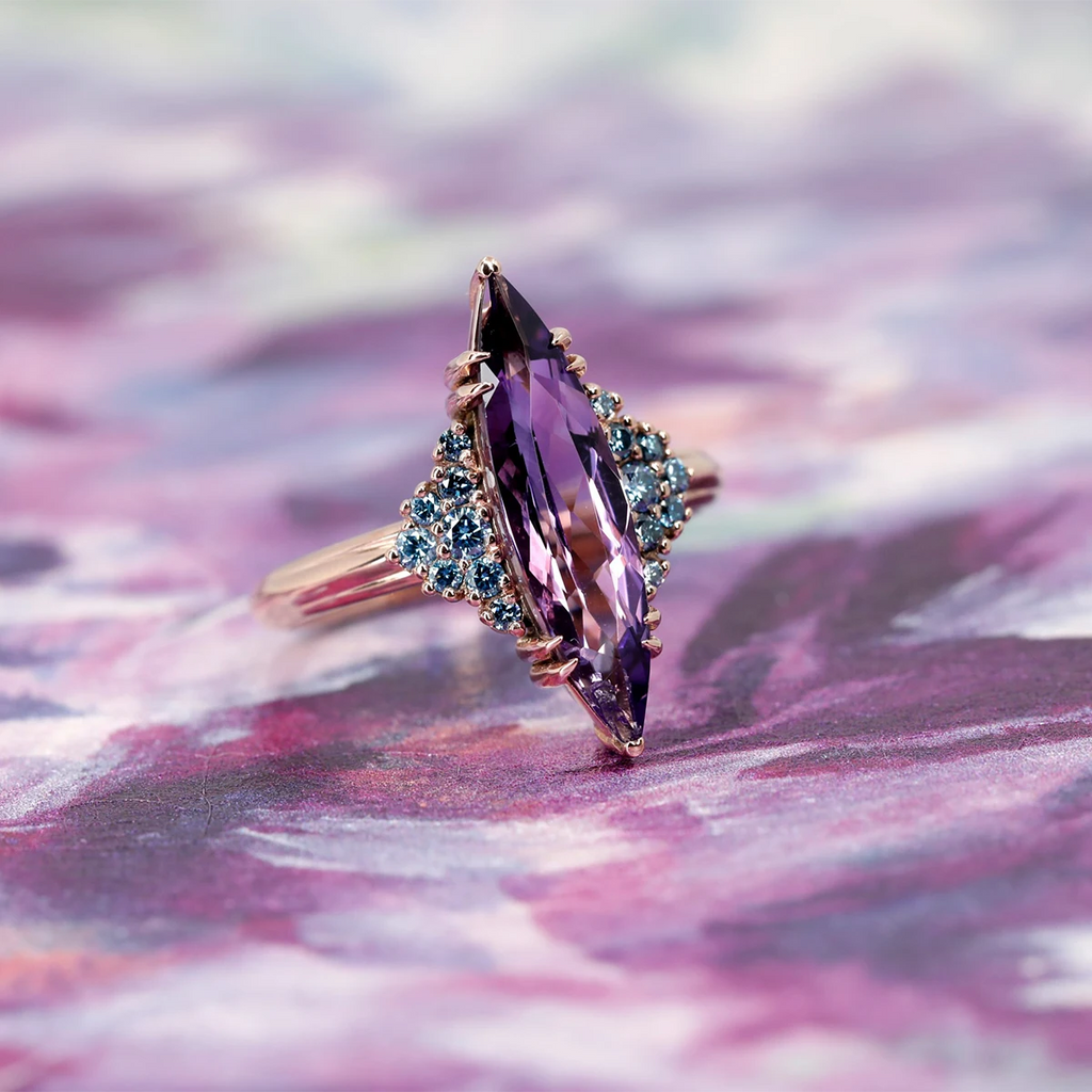 Statement marquise amethyst ring with vivid blue diamond side stones, set in rose gold, displayed on a purple painted background – Bena Jewelry Montreal.