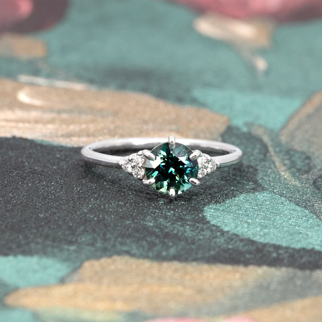 Front view of a round teal sapphire ring with white diamond accents, set in 14k white gold, placed on a soft floral background. Fine jewelry by Maksym Jewelry in Montreal, available at Ruby Mardi.
