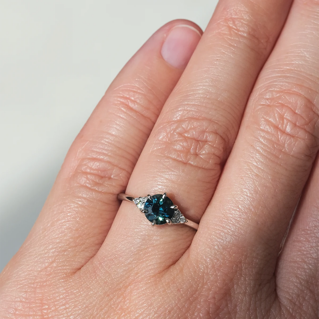 Teal sapphire engagement ring with white diamond accents in 14k white gold, worn on hand. Handmade in Montreal by Maksym Jewelry, available at Ruby Mardi.
