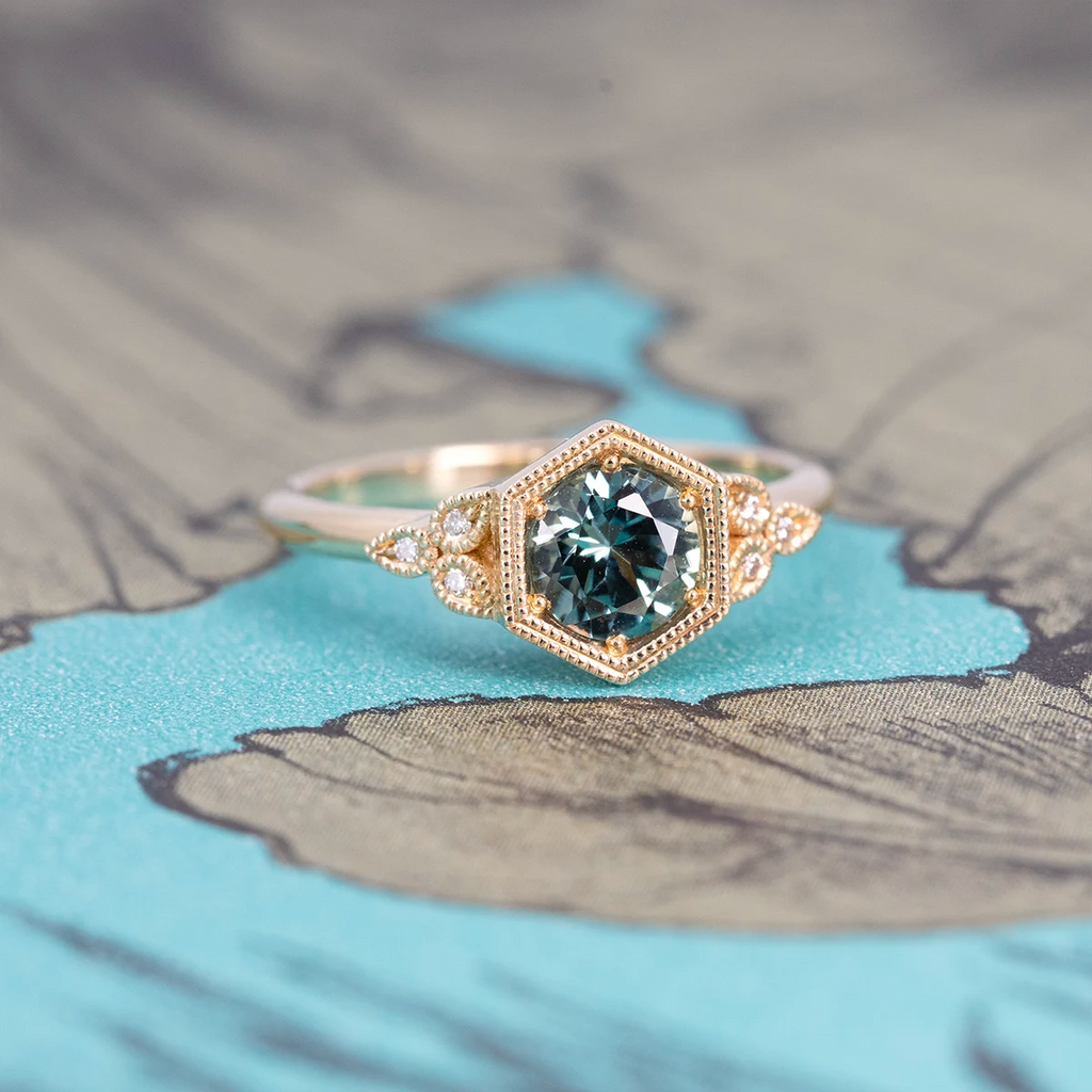 Teal sapphire engagement ring with hexagonal vintage-inspired bezel and diamond accents in yellow gold, displayed on artistic background, designed by Emily Gill, available at Ruby Mardi, Montreal Canada.