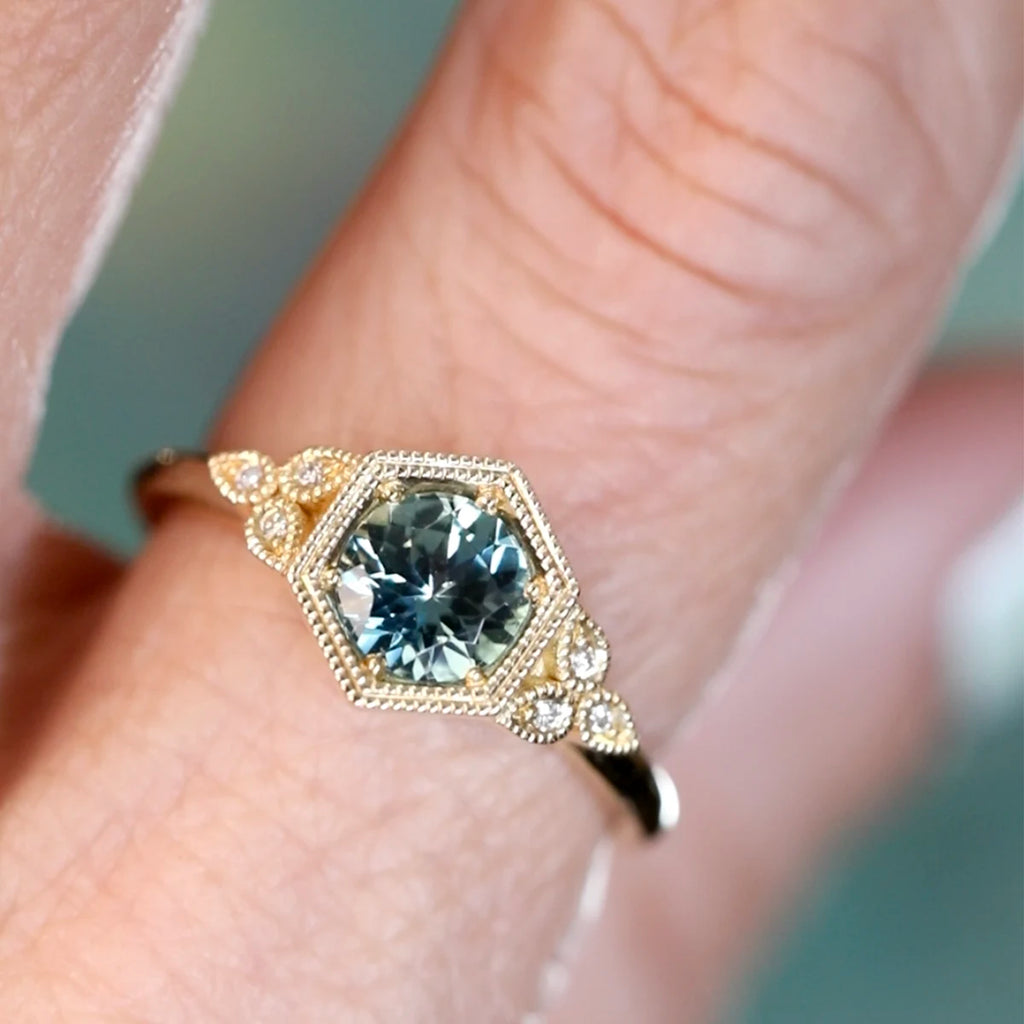 Teal sapphire engagement ring with hexagonal vintage bezel and diamond accents in yellow gold, worn on hand, designed by Emily Gill, available at Ruby Mardi, Montreal Canada.