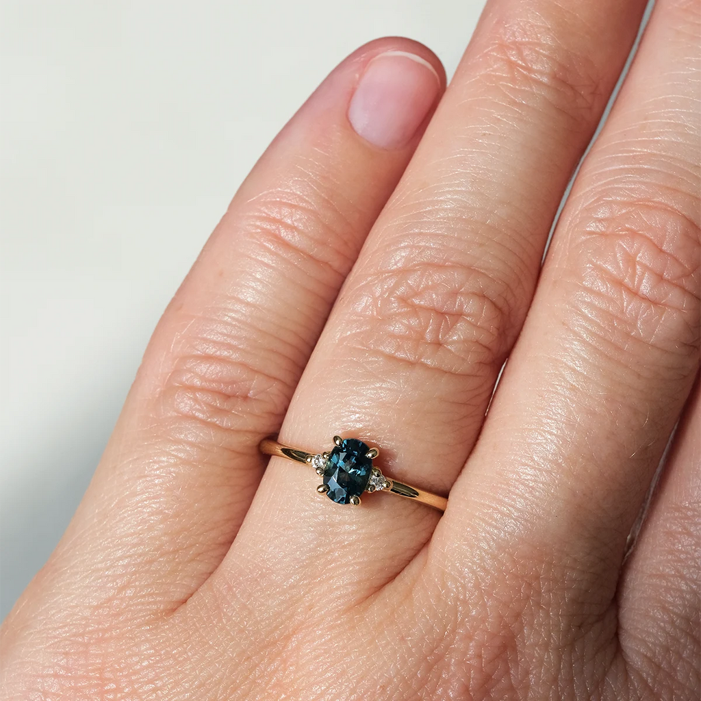Oval teal sapphire engagement ring with side diamonds, set in yellow gold, worn on hand. Handmade in Montreal by Maksym Jewelry. Available at Ruby Mardi, Canadian designer jewelry store.