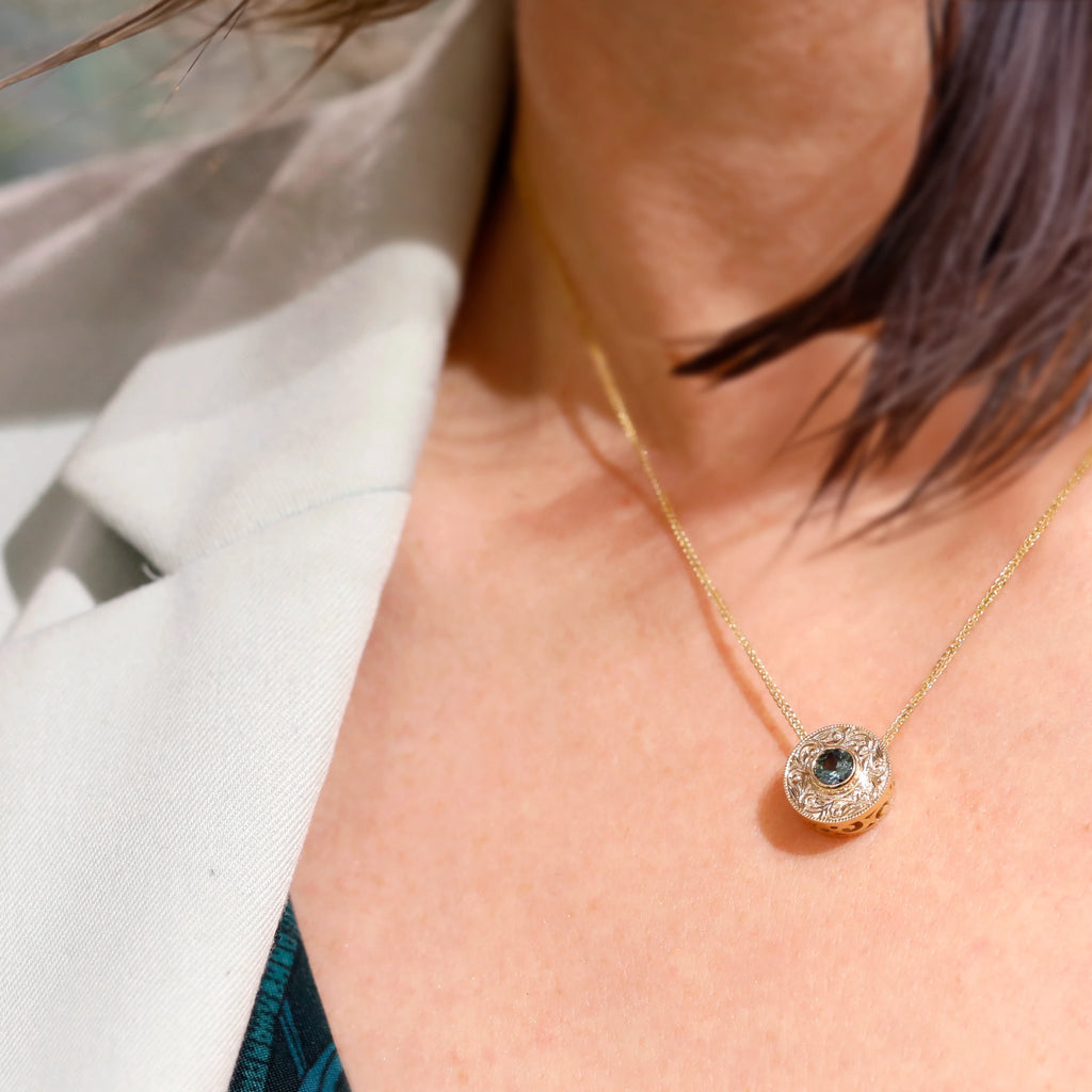 This woman is wearing Deborah Lavery's pendant with a round-shaped, teal-colored sapphire. Mounted in yellow gold and engraved by hand, this fine jewel is a unique creation made in Canada and on sale at the only jewelry gallery in Montreal, the Ruby Mardi jewelry store, specialist in one-of-a-kind jewelry made by independent jewelers.