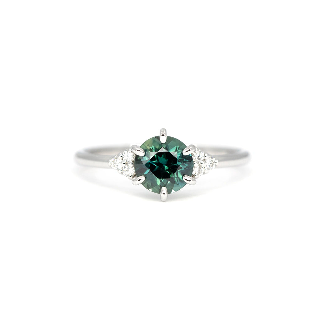Centered shot of a teal green sapphire engagement ring with pear-shaped and round diamonds on each side, crafted in white gold by Maksym Jewelry, Ruby Mardi, Montreal.