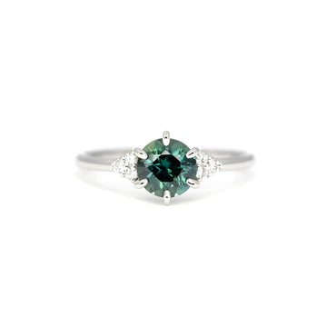Centered shot of a teal green sapphire engagement ring with pear-shaped and round diamonds on each side, crafted in white gold by Maksym Jewelry, Ruby Mardi, Montreal.