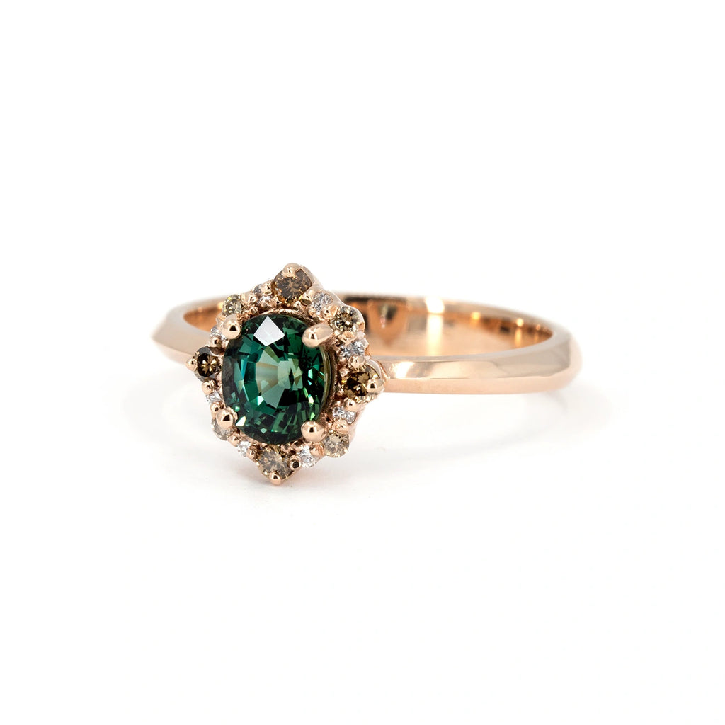 Close-up of a rose gold engagement ring featuring an oval teal sapphire, surrounded by a pointed halo of natural brown and white diamonds. Handcrafted by Ruby Mardi in Montreal.