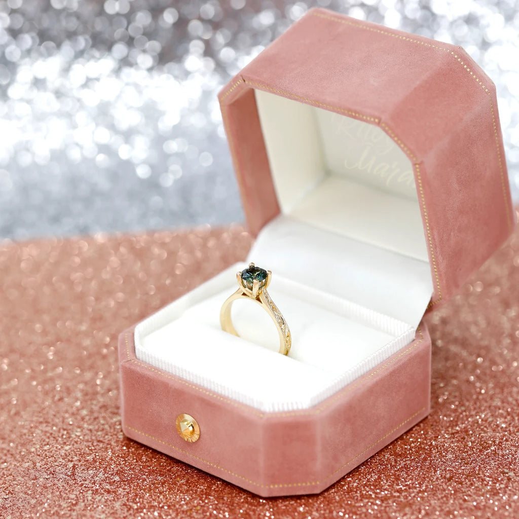 Solitaire engagement ring with a teal sapphire and scroll engravings on the gold band, seen on its gift box. This engagement ring was handmade in Canada by Deborah Lavery and is available exclusively at Ruby Mardi in Montreal.
