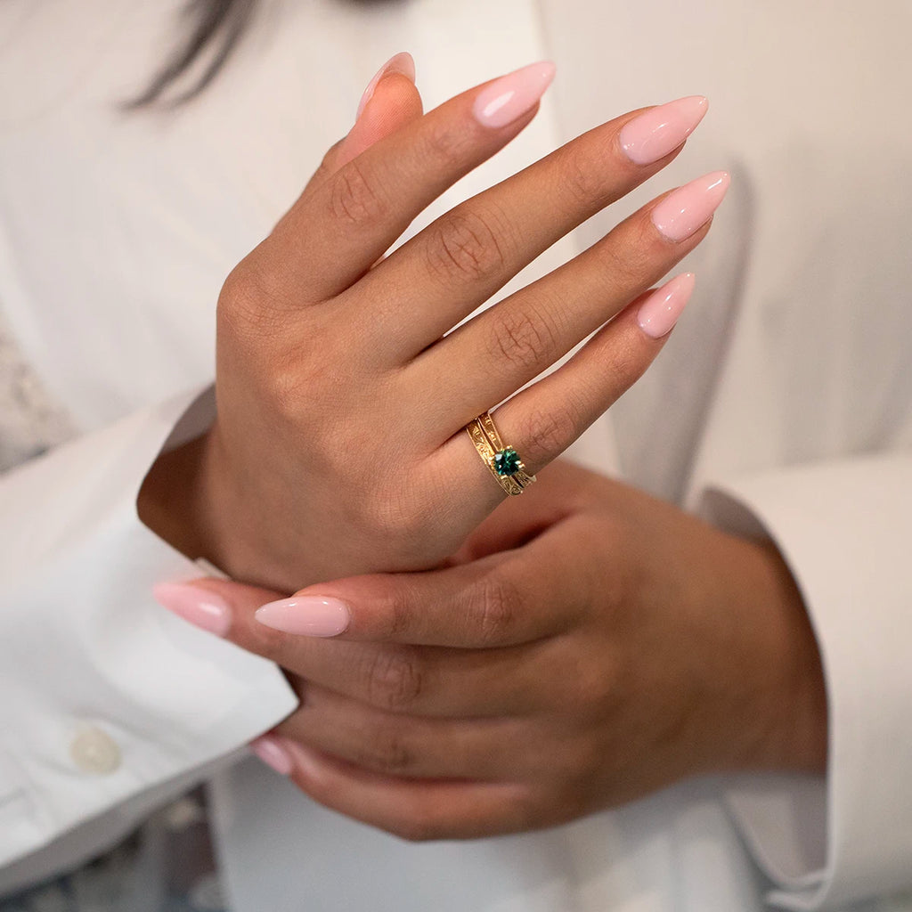 We see two hands of a woman wearing a white shirt with long pink nails wearing two rings stacked together on her pinky finger. These are two handmade creations by Toronto jewelry designer Deborah Lavery. They are a solitaire engagement ring with an Australian green sapphire, and a yellow gold wedding band with handmade scroll engravings. Fine jewelry available at Ruby Mardi.