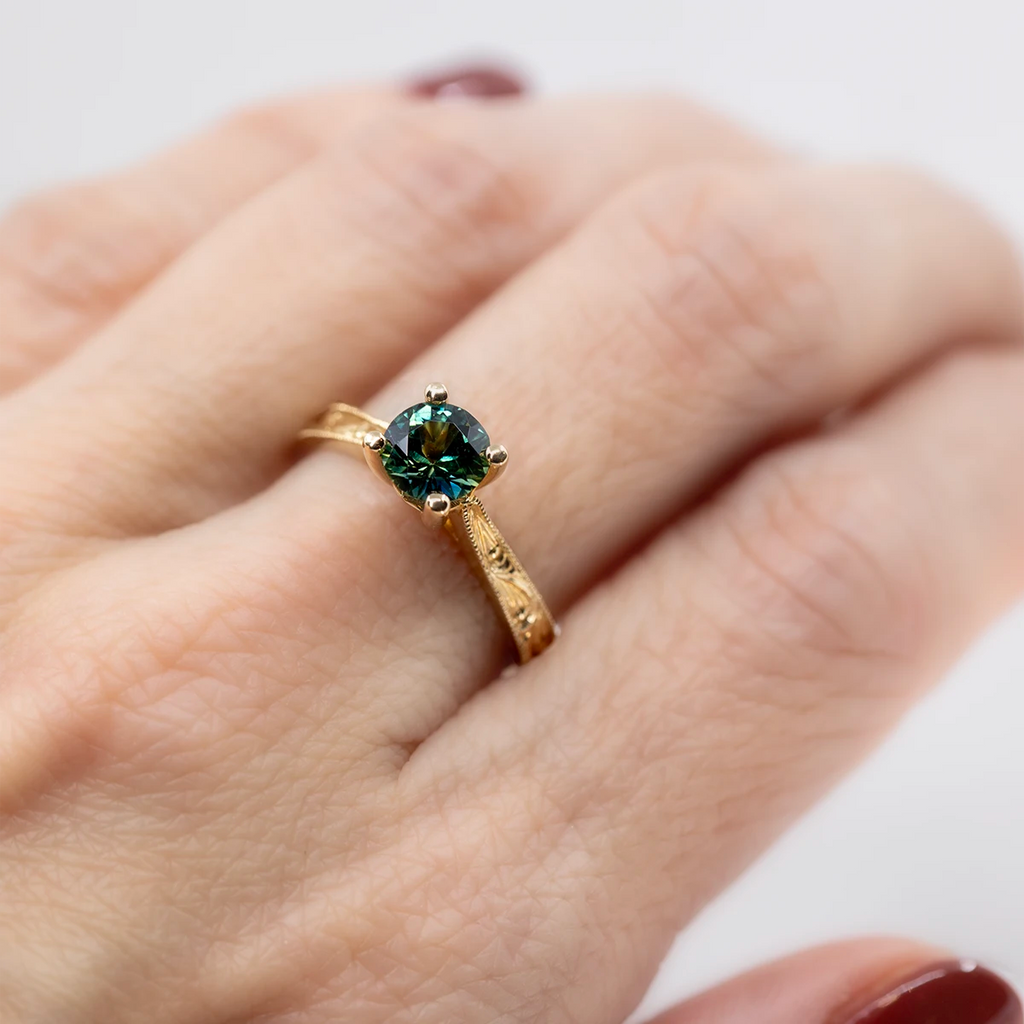 Close-up of an engagement ring worn on a hand with dark red nails. The yellow gold ring features a round teal sapphire, raised in a classic fashion to easily accommodate a wedding band. The ring features hand-engraved scrolls. This bridal jewel was entirely handcrafted by Deborah Lavery. Ruby Mardi represents this jewelry artist, as well as several other talented independent jewellery designers.