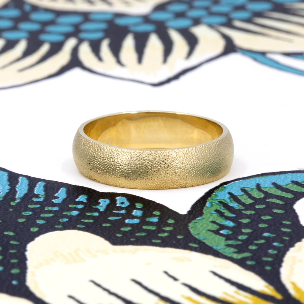 Handcrafted textured gold wedding band with a soft satin finish, displayed on an artistic floral background. Available at Ruby Mardi, Montreal.