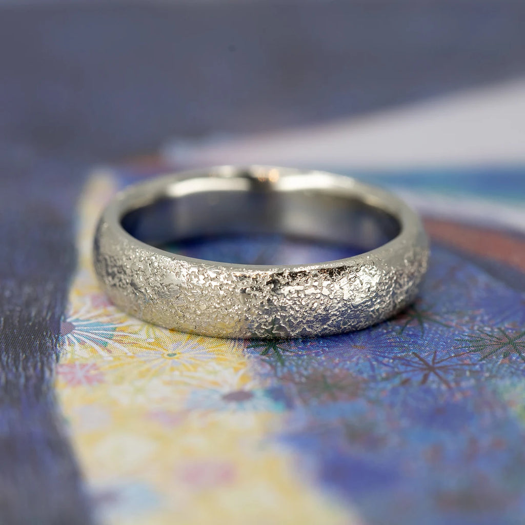Close-up of a textured white gold wedding band with a domed profile, handcrafted by Fluid Jewellery, resting on a colorful patterned background.