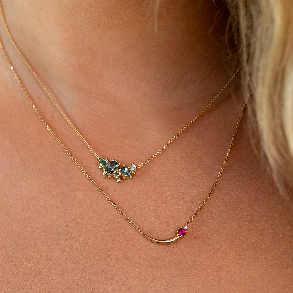Close-up of layered gold necklaces: the upper one featuring a cluster of blue-green tourmalines and diamonds, the lower one a curved pendant with a vivid pink gemstone. Fine jewelry from Ruby Mardi, Montreal.