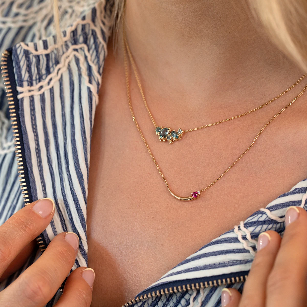 ChatGPT said:

Layered necklaces styled with a striped blouse: the upper featuring teal tourmalines and diamonds, the lower a gold curved pendant with a pink gemstone. Fine designer jewelry from Ruby Mardi, Montreal.