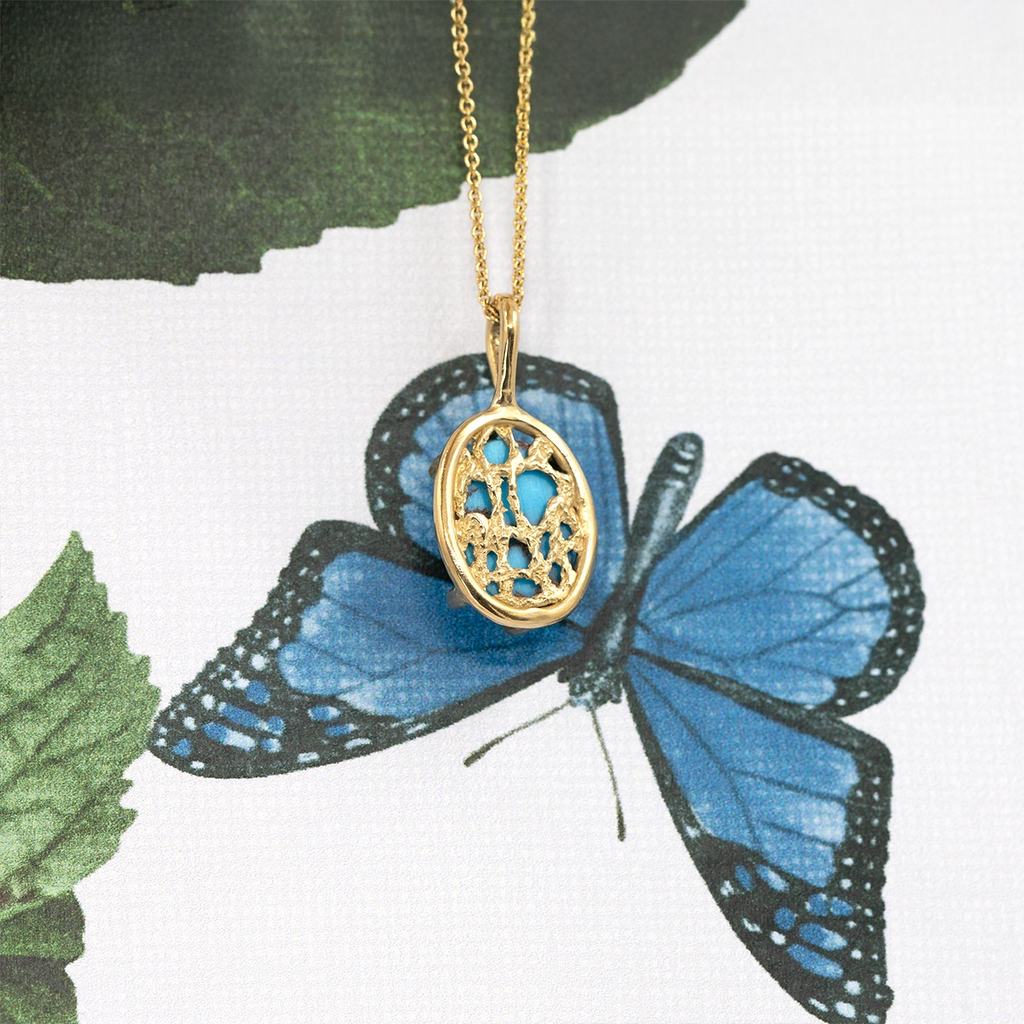 Handcrafted gold pendant by Ruby Mardi, a Montreal-based boutique featuring Canadian jewelry designers. The pendant showcases an openwork lace design with a natural turquoise gemstone at its center, hanging from a delicate gold chain. Photographed on a blue butterfly background, the image highlights the artistry and organic elegance of Ruby Mardi’s fine jewelry collection.