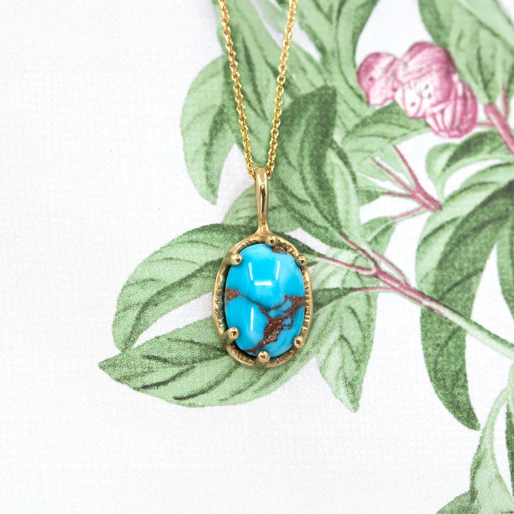 Handcrafted turquoise pendant by Ruby Mardi, a Montreal boutique showcasing Canadian jewelry designers. The oval turquoise gemstone, featuring natural brown veining, is set in an engraved gold bezel and suspended from a fine gold chain. Photographed against a botanical background, highlighting the artisanal craftsmanship and organic beauty of Ruby Mardi’s designer jewelry.