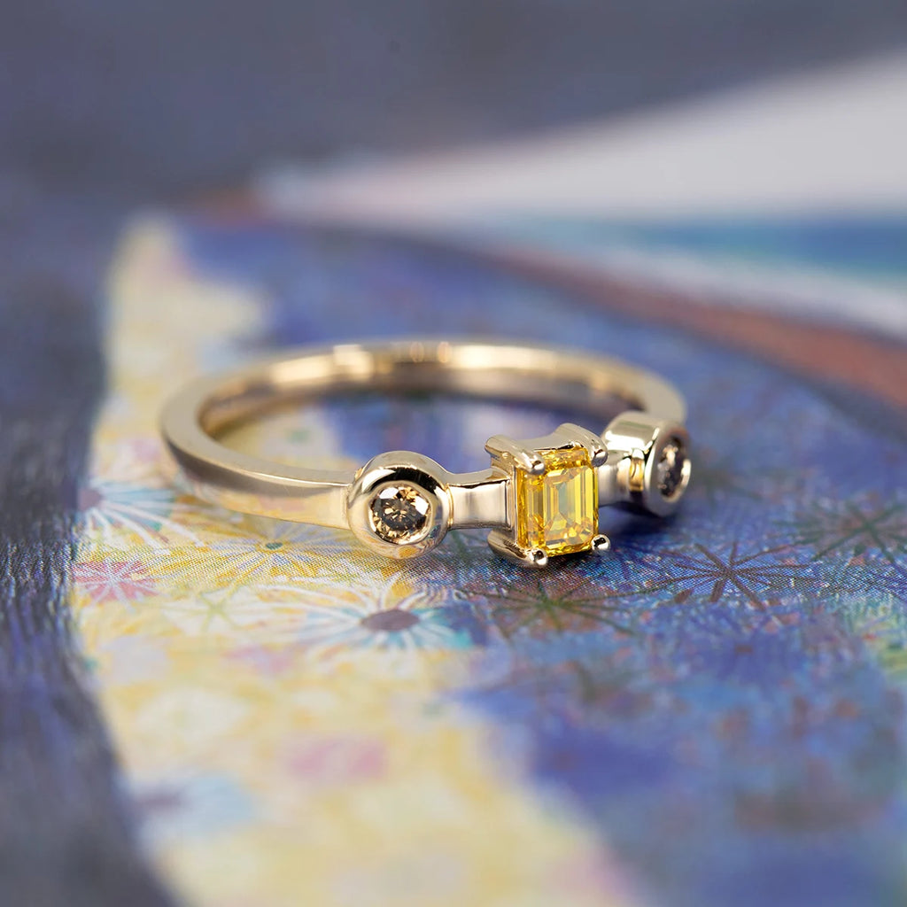 Yellow gold trilogy ring with an emerald-cut yellow diamond and two round natural brown diamonds in bezel settings, displayed on a colorful textured background, designer jewelry available at Ruby Mardi in Montreal.
