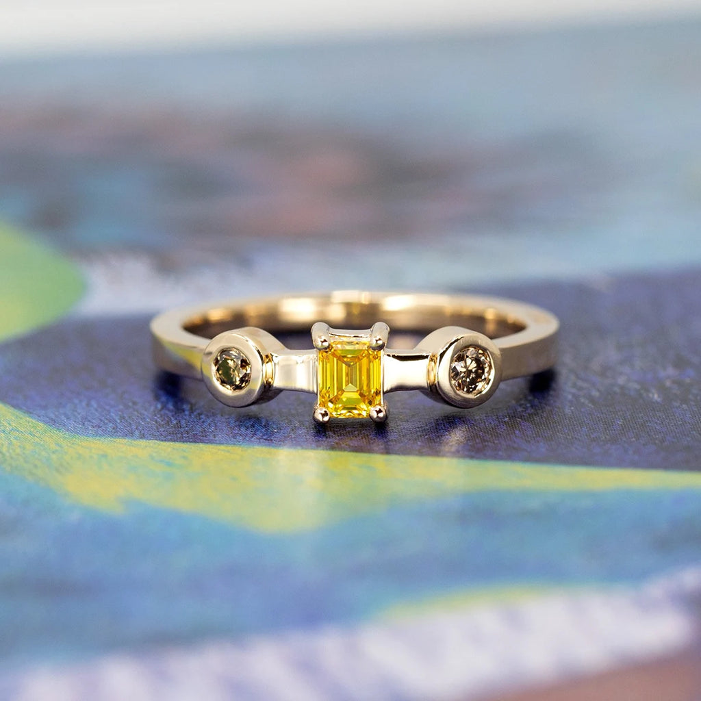 Yellow gold trilogy ring with an emerald-cut yellow diamond and two bezel-set brown diamonds, photographed on a colorful artistic background, contemporary designer jewelry from Ruby Mardi, Montreal.

