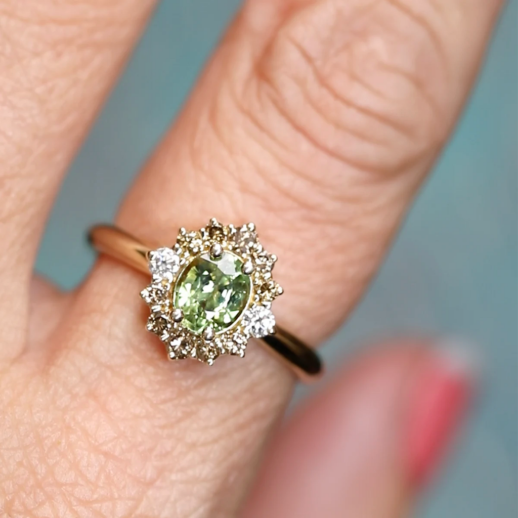 Close-up of a yellow gold engagement ring worn on a finger, featuring an oval green sapphire framed by a halo of champagne diamonds and accented with two white diamonds, handcrafted by Ruby Mardi Jewelry in Montreal.