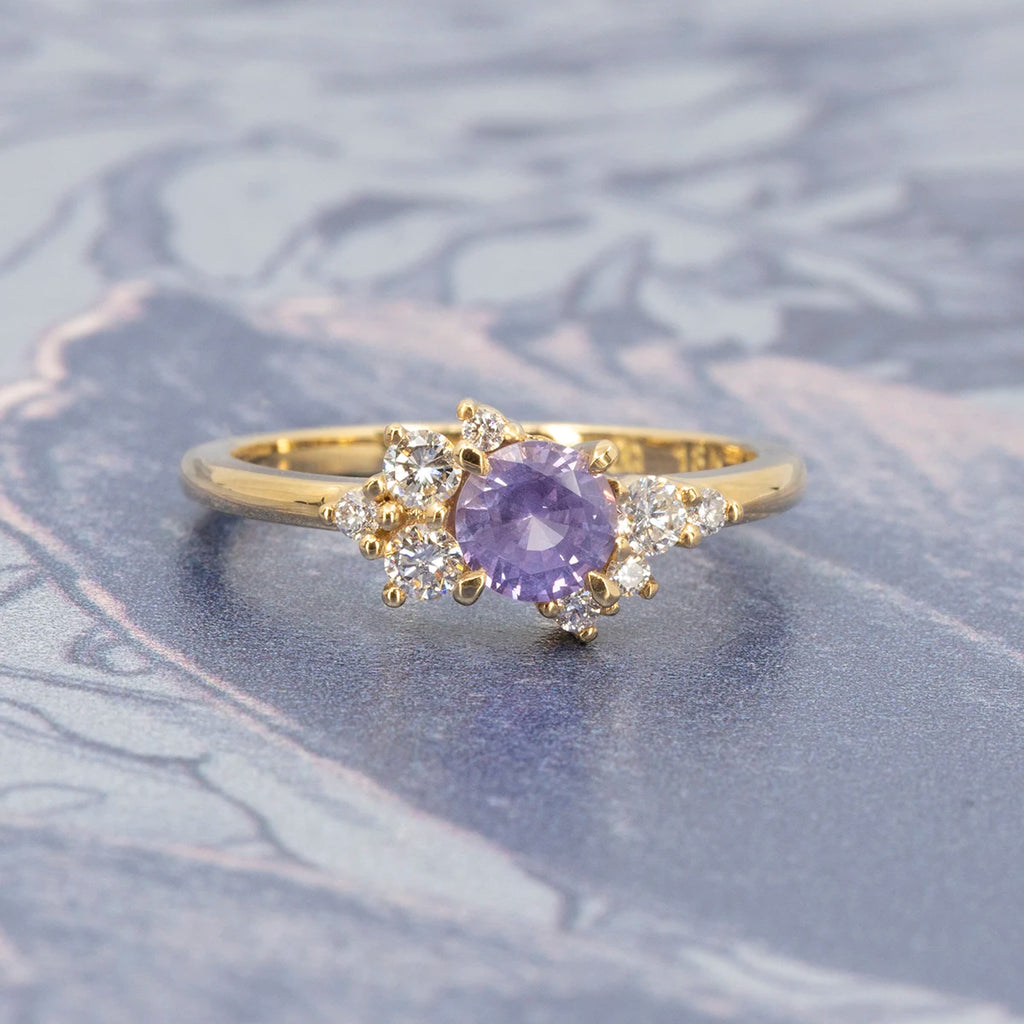 A gold ring designed by Justine Quintal with a round purple sapphire center and asymmetrical clusters of small round white diamonds, shown on a soft marbled blue background.