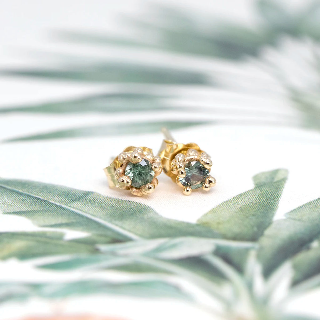 Pair of one-of-a-kind teal sapphire stud earrings handcrafted in 14k yellow gold by Meg Lizabet. Fine organic jewelry available at Ruby Mardi, Montreal, Canada, shown on a botanical background.