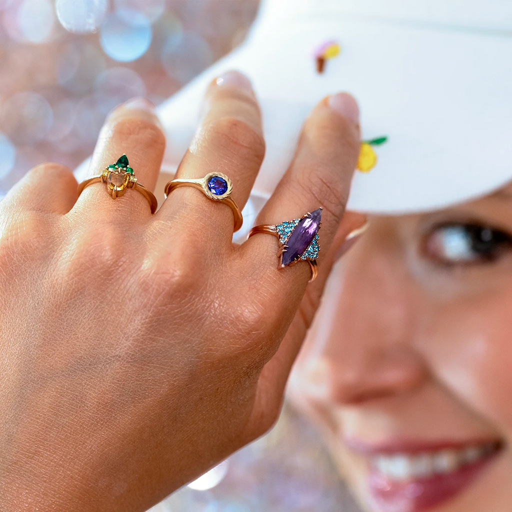 Close-up of a smiling woman wearing three unique rings on her hand while holding the brim of a white cap. The rings include a triangular green gemstone ring, a round blue gemstone ring in a bezel setting, and a striking marquise-cut purple gemstone ring accented with small blue stones, all set in yellow or rose gold. Fine jewelry by Ruby Mardi, handcrafted in Canada.