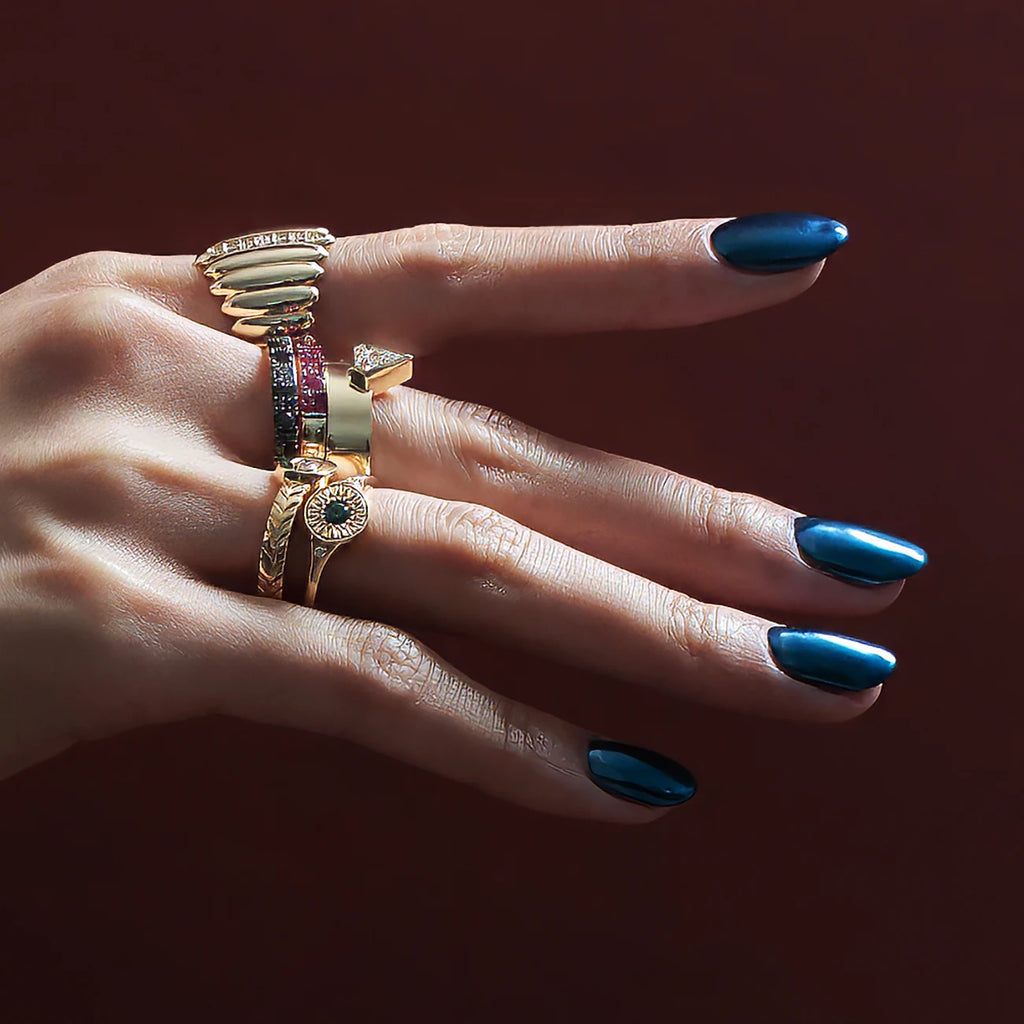 A hand in close shot studio photography is modelling for fine jewelry pieces. The hand has dark blue metallic nails. The background is red wine color. The hand wears 6 unique designer rings of unique different styles. All of them are in yellow gold. Gemstones seen are diamonds, white sapphire, green sapphire, natural black diamonds and rubies.