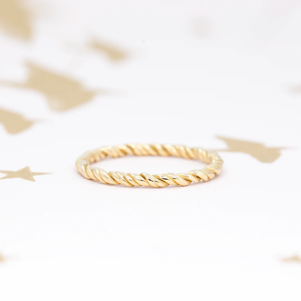 Twisted gold ring displayed on a white surface with gold stars. The rope-style band shines with a subtle glow, part of Ruby Mardi’s curated indie jewelry collection, made in Montreal.