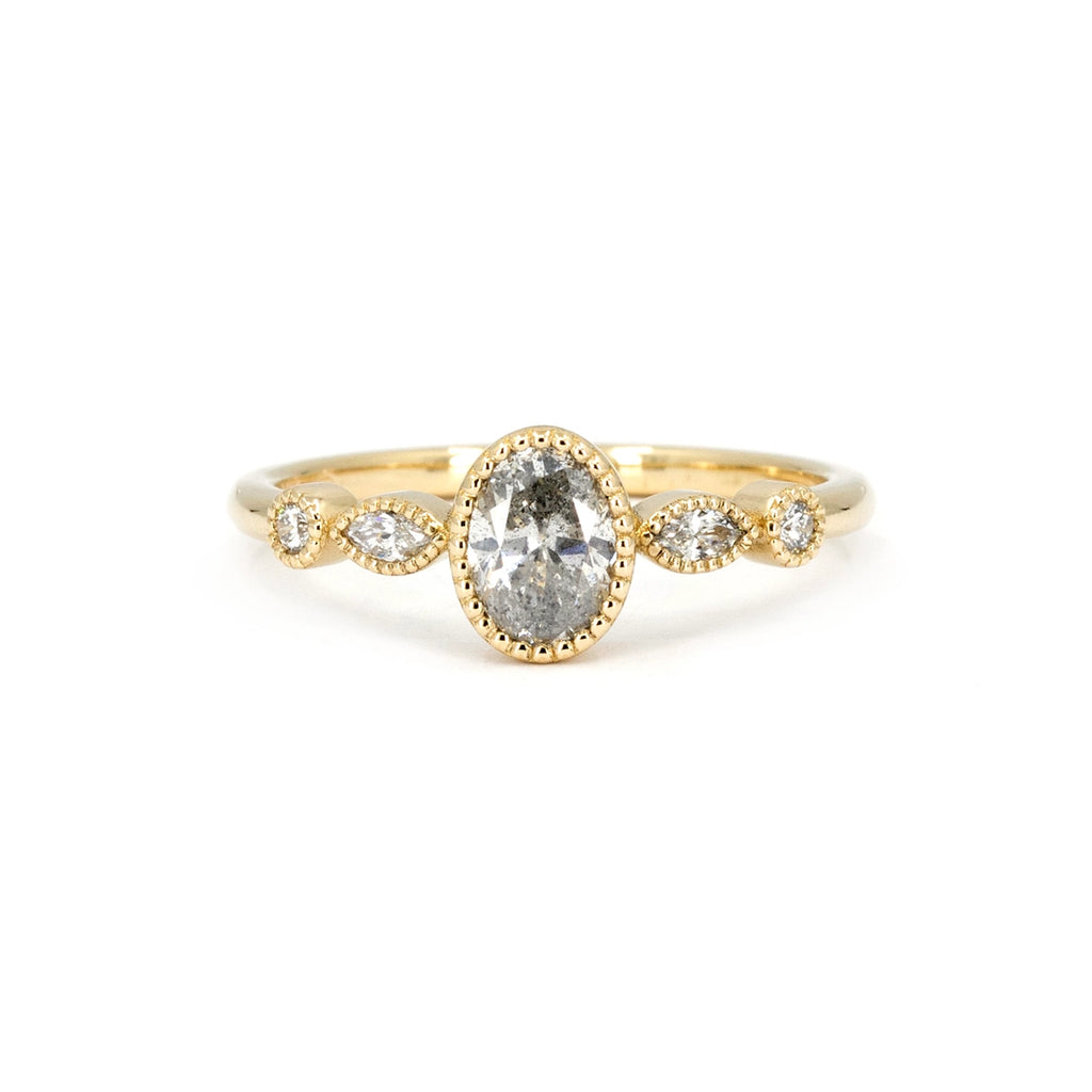 An engagement ring with vintage charm is photographed against a white background. The ring is in ethical gold (Fairmined certified) and features 5 diamonds all bezel set with miligrain details. The central natural diamond is oval-shaped and is salt-and-pepper. The side diamonds are lab grown. This fine jewel is a creation of Canadian brand Fluid Jewellery. This wedding ring is available at Ruby Mardi, a fine luxury jewelry store.