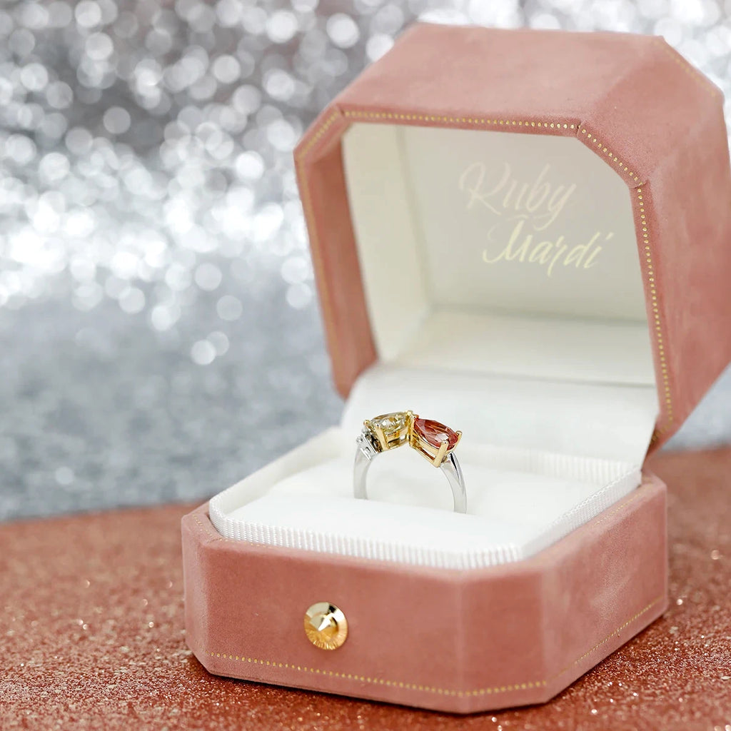 A toi&moi designer engagement ring featuring a natural yellow diamond and a peach spinal is seen photographed in the gift box of the jewelry store Ruby Mardi, a jewellery gallery that carries more than 20 independent jewelry brands. The box is in pink velvet and is seen on a background made of pink and silver glitters.