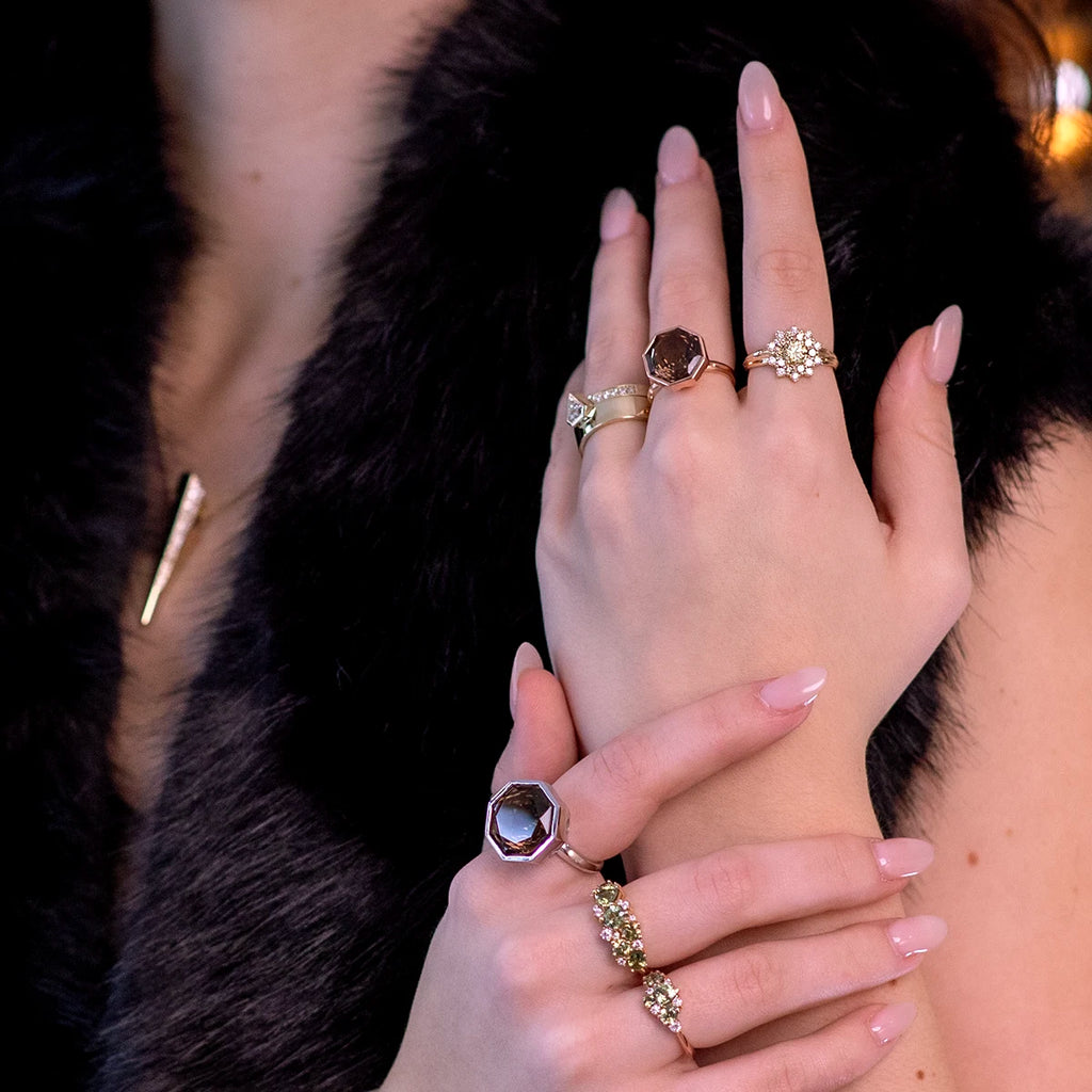 A close-up image showcasing a woman's hands adorned with multiple elegant rings in various designs. The rings feature unique gemstones, including a large hexagonal gem, clusters of golden and white diamonds, and intricate gold settings. Her nails are manicured in a soft nude color, and she is dressed in a luxurious black fur coat, adding a touch of sophistication. A gold pendant necklace is subtly visible in the background.