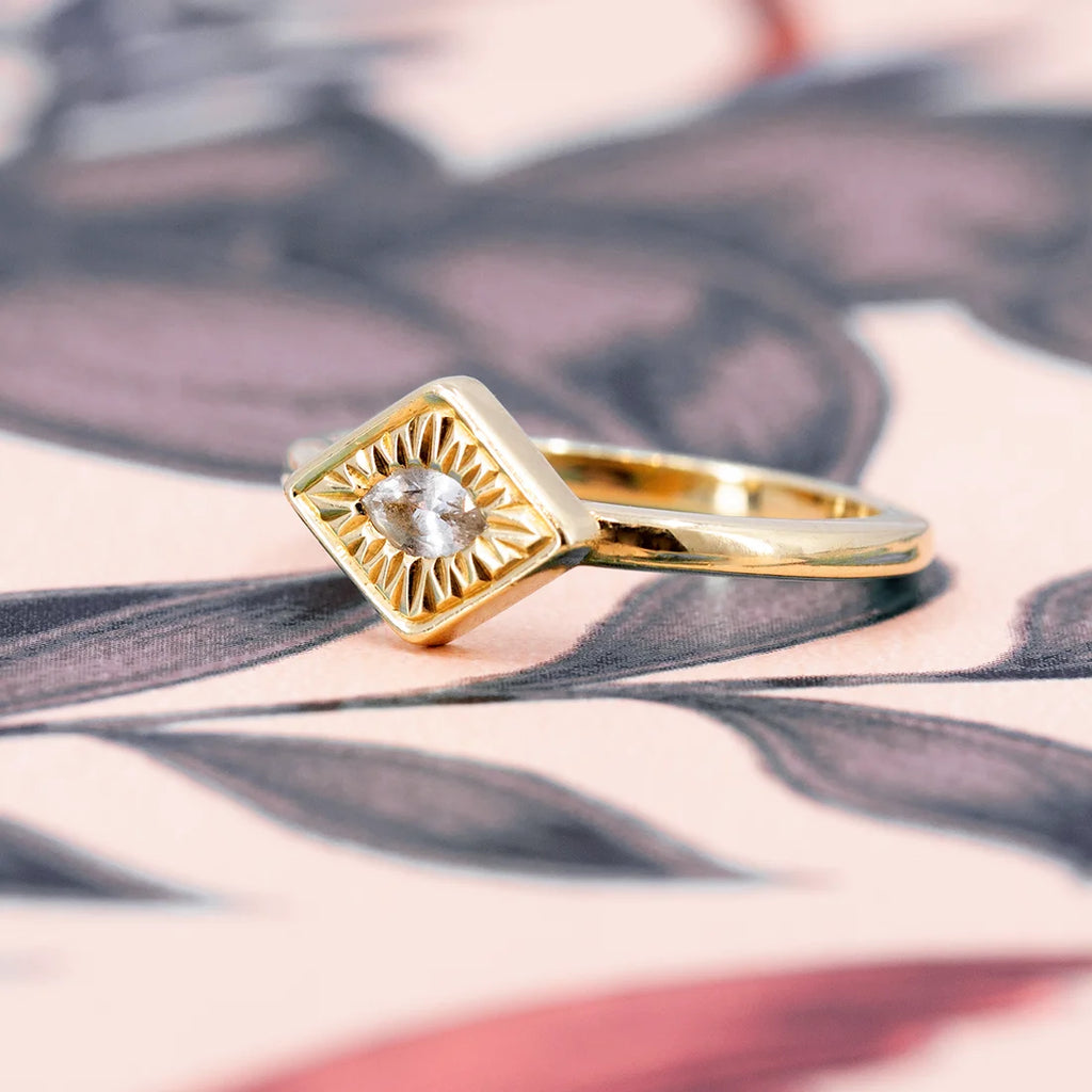Close up on a signet engagement ring : thick gold, hand engraved details, white sapphire. Photographed on an abstract background. Find this designer piece from Savannah Jones at Ruby Mardi, an indie Canadian jewelry store that curate the work of talented emerging and established designers.