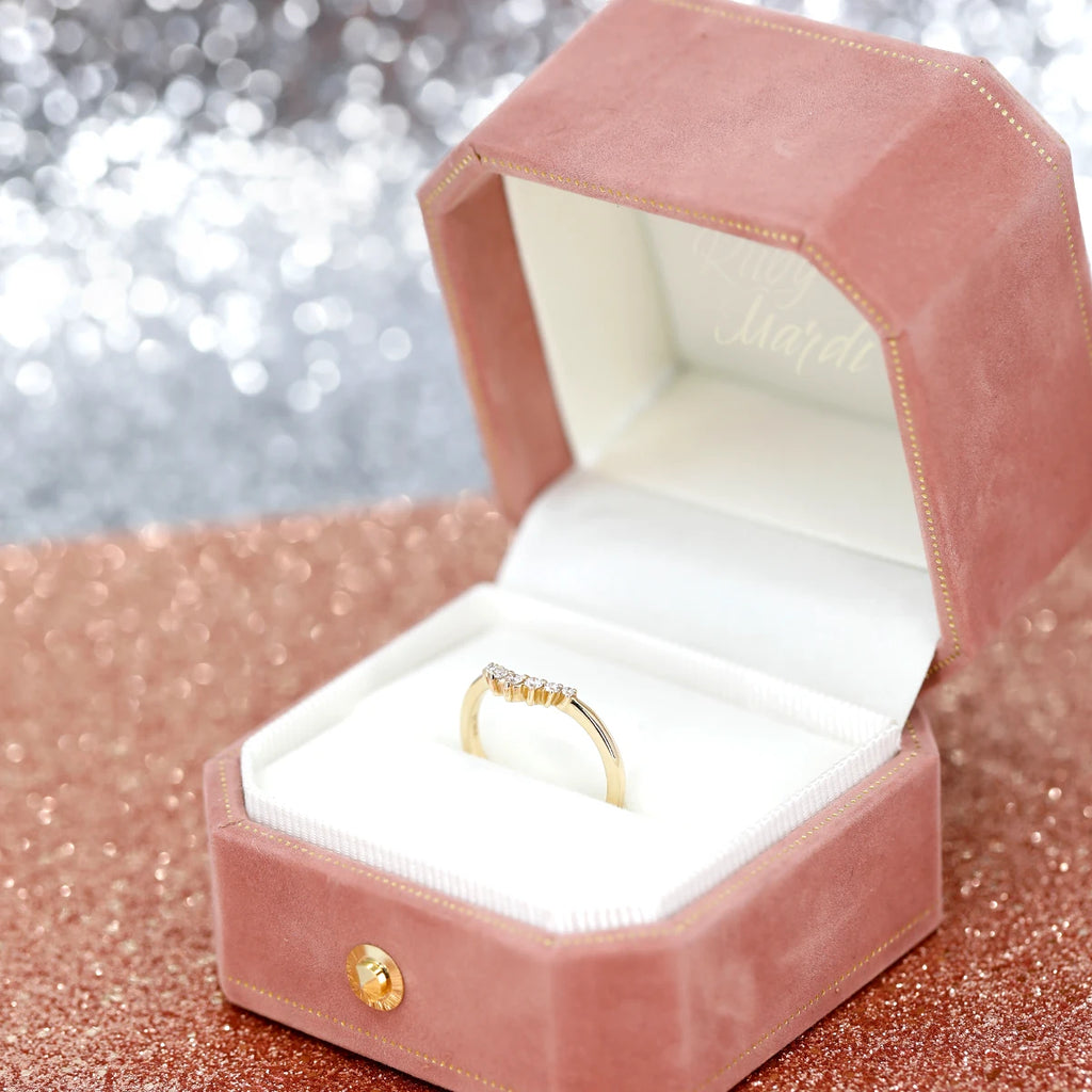 A yellow gold wedding band with five small round brilliant-cut white diamonds is photographed in its pink velvet gift box. The box rests on a background of pink sequins. 