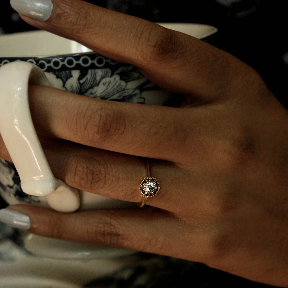 girl's hand wearing a white sapphire black diamond halo bridal engagement ring made in montreal by Liane Vaz finest jewellery designer at best jewelry store in montreal ruby mardi on a dark holding a tea cup background